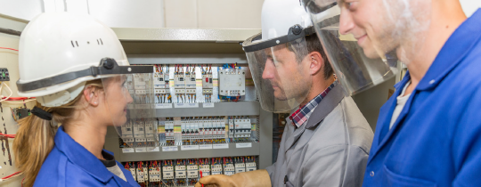 3 coworkers where work helmets and face shields looking at electrical panel